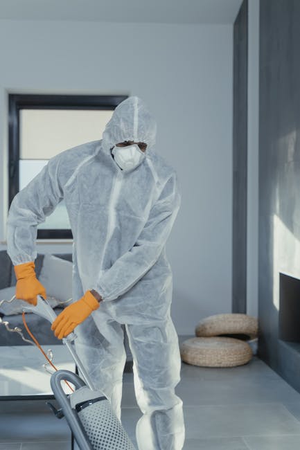 A professional cleaner dressed in a full white protective suit, face mask, and orange gloves is operating a vacuum cleaner during a deep surface cleaning process in a modern, well-lit room. The space