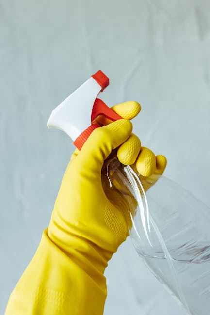 A professional cleaner dressed in a full white protective suit, face mask, and orange gloves is operating a vacuum cleaner during a deep surface cleaning process in a modern, well-lit room. The space