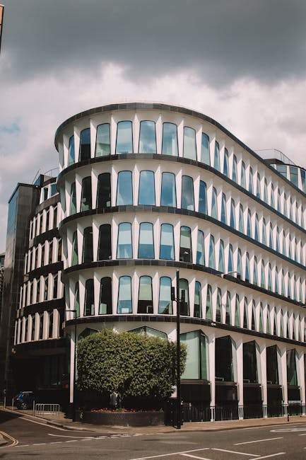 Exterior view of a modern multi-story office building with curved architecture and large glass windows on each floor, reflecting the cloudy sky above. The building's facade features white vertical str