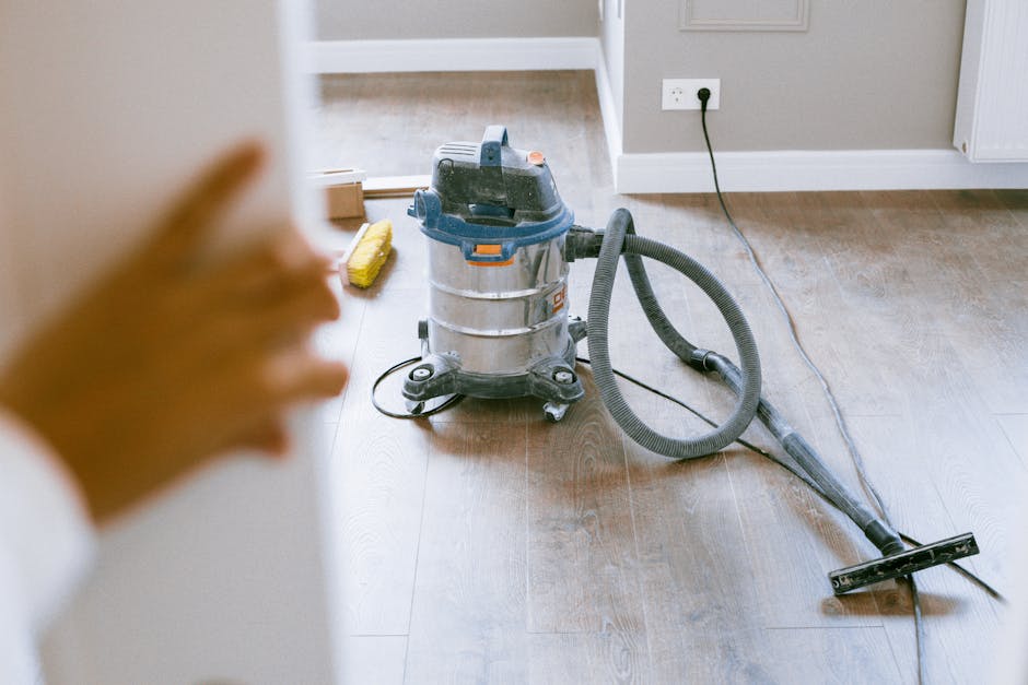 A woman performs surface cleaning in a modern kitchen, wearing a grey t-shirt and red overalls. She is using a mop to clean the tiled floor near a white wall, with a small blue bucket placed beside he